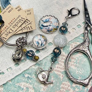 May include: An assortment of craft supplies including embroidery floss, a key ring with charms, decorative scissors, and beaded accents. The charms feature teacups and teapots. The floss is in shades of blue and green.