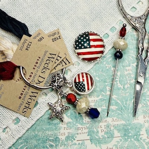 Puede incluir: Una colección de suministros para manualidades que incluye hilo Weeks Dye Works, un pin patriótico y un dije decorativo con detalles de estrellas y cuentas. También se incluyen una aguja, unas tijeras y un pequeño pin de la bandera estadounidense.