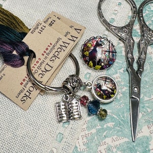 May include: A silver key chain with a book charm, two stained glass charms, and colorful beads. The key chain is next to a pair of silver scissors and a tag that reads "VW Weeks Dye Works" with the text "Specializing in Hand Dyed Fibers" and "FLOSS COLLECTION".