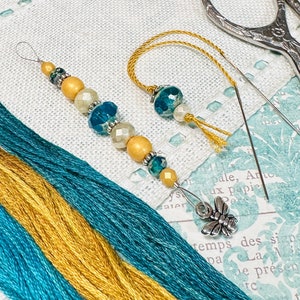 May include: A close-up of a needle threader with a beaded tassel, a needle, and a pair of scissors. The threader has a gold and teal color scheme. The needle is silver and the scissors are silver with a floral design. The background is a white fabric with a floral pattern.