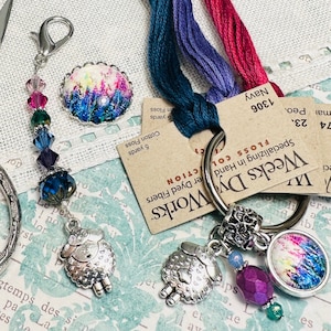 May include: A set of two needle minders with colorful floral designs. Each minder is attached to a silver charm with a sheep design. The minders are attached to a silver ring with a beaded chain and a pair of scissors. The image also includes two floss cards with the text "Weeks Dye Works" and "Specializing in Hand Dyed Fibers".