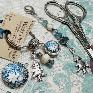 May include: A silver embroidery scissor with a decorative handle, a silver key ring with a blue and white snowflake charm, a blue and white star charm, a silver tree charm, and a white bead charm. The key ring is attached to a tag that reads "Weeks Dye Works, Specializing in Hand Dyed Fibers, Floss Collection".