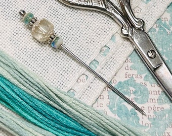 May include: A close-up of a needle with a clear glass bead and silver accents, resting on a white linen surface.  A pair of silver scissors is also visible in the background.