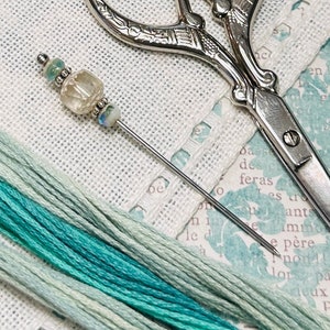 May include: A close-up of a needle with a clear glass bead and silver accents, resting on a white linen surface.  A pair of silver scissors is also visible in the background.