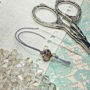 May include: A pair of silver-toned metal embroidery scissors with floral engravings, a long sewing needle threaded with lavender cord, a purple and yellow patterned bead, a small green bead, and a pile of clear faceted crystal beads.
