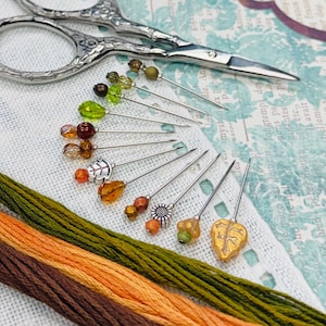 May include: A set of decorative sewing pins with various bead and metal leaf-shaped heads in shades of green, orange, and brown, arranged in a fan pattern alongside a pair of silver-toned metal embroidery scissors and three strands of embroidery floss.