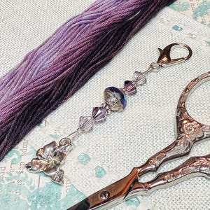 Puede incluir: Una tijera de plata con un diseño adornado y un hilo de bordar morado y gris con un dije de cuentas de plata.