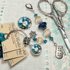 May include: A collection of craft supplies including embroidery floss, scissors, and decorative charms. The floss is teal and green. The charms feature a snowman design. The scissors are silver. The image includes tags that read "Week Dye Works".