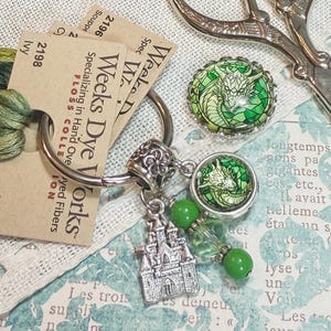 May include: A silver key ring with a silver castle charm, two green glass dragon charms, and green glass beads. The key ring is attached to two cards with the text "Weeks Dye Works" and "Specializing in Hand Oveyed Fibers".