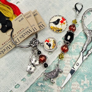May include: An assortment of craft supplies including embroidery floss, a scissor, and decorative charms. The floss is black, yellow, and red. Charms feature bird and floral designs. The scissors are silver-toned.
