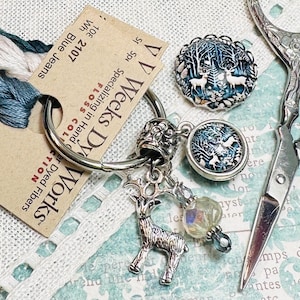 May include: A silver key chain with a charm of a deer, a glass bead, and two round glass charms with a winter forest scene. The charms are attached to a silver ring with a silver bead. The key chain is on a white fabric background with a tag that reads "VV Weeks Dy Works" and "Specializing in Hand Dyed Fibers".