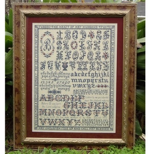 May include: A framed cross-stitch sampler with a cream-coloured background and red border. The sampler features a complete alphabet in various fonts, numbers, and a verse: "Before I was born the Lord called me by name. He said to me, "You are my servant. I have chosen you and called you by name."