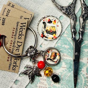 Peut inclure: Un assortiment de fournitures d'artisanat comprenant des cartes de fil Weeks Dye Works, des ciseaux argentés et un charm avec un motif de chat. Le charm a un accent de perles rouges, jaunes et noires, et un charm en forme de feuille d'argent. Les cartes de fil portent le texte "Weeks Dye Works" et "Liberty".