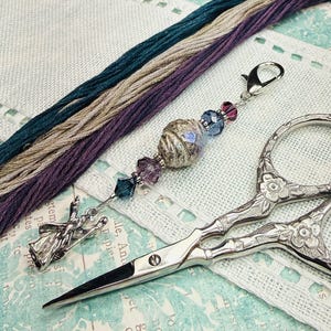 May include: A silver-toned metal scissor fob featuring a central textured bead, faceted blue and purple crystal beads, and a silver-toned metal charm shaped like a witch on a broomstick, attached to a lobster claw clasp for easy attachment.