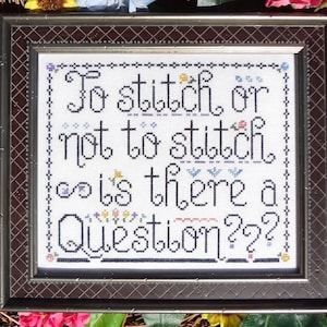 Puede incluir: Un punto de cruz enmarcado con el texto "To stitch or not to stitch, is there a question??" en hilo negro sobre tela blanca. El punto de cruz está decorado con bordados florales de colores.