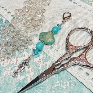 Puede incluir: Un par de tijeras decorativas de plata con un charm de cuentas turquesa y oro. El charm presenta un caballito de mar y una cuenta turquesa y oro. Las tijeras están sobre un fondo de tela blanca.