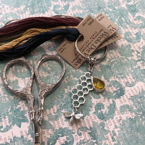 May include: A pair of silver embroidery scissors with a honeycomb charm and a yellow glass bead. The charm is attached to a silver key ring with a tag that reads "Weeks Dye Works Specializing in Hand Overed Fibers 1103 Pea Coat 1270 Rum F 2103".