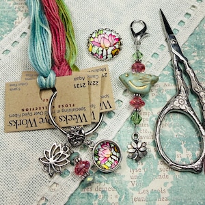 May include: An assortment of craft supplies including embroidery floss, a charm bracelet with a lotus flower pendant, a bird-shaped bead, and decorative scissors. The floss is in shades of teal, pink, and green. The scissors have a floral design.