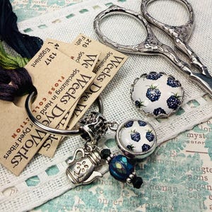May include: An assortment of craft supplies, including embroidery floss, scissors, and decorative charms. The floss is a blend of green, purple, and blue. The scissors are silver with ornate handles. Charms feature a teapot and a blackberry design.