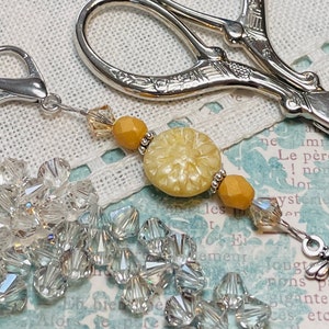 Puede incluir: Un charm de tijera plateado con un botón amarillo, cuentas transparentes y ámbar, y un charm de abeja plateado. El charm está unido a un clip plateado.