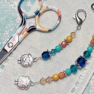 May include: Two silver sheep charms attached to beaded stitch markers. The stitch markers are made with turquoise, blue, and yellow beads. The stitch markers are attached to silver clasps. A pair of colorful scissors are also in the image.