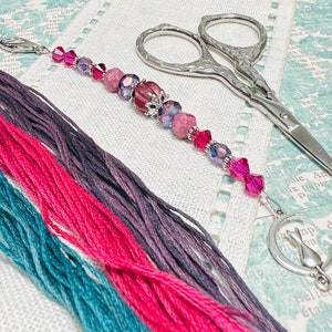 May include: A beaded scissor fob with pink, purple, and clear beads, a silver cat charm, and a pair of silver embroidery scissors. Colorful embroidery floss in teal, pink, and purple is also visible.