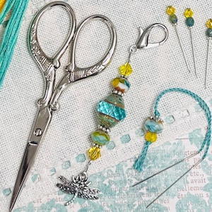 May include: A turquoise and yellow beaded needle minder with a dragonfly charm, a pair of silver scissors, and several needles with turquoise and yellow beads.