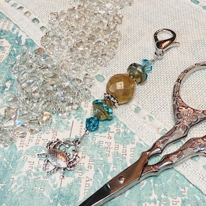 May include: A silver pair of decorative scissors with a beaded charm attached. The charm features clear and blue glass beads, a brown stone, and a silver crab.