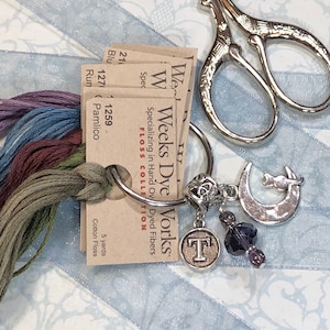 May include: A close-up of a set of embroidery floss spools in a variety of colours, including purple, blue, brown, and green. The spools are labelled with the brand "Weeks Dye Works" and the colour numbers. The spools are attached to a silver keyring with a charm in the shape of a crescent moon and a cat.