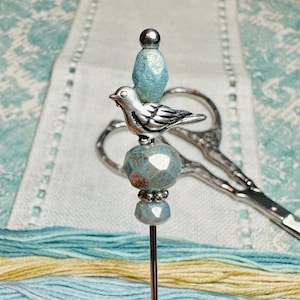 May include: A silver bird charm sits atop a needle with blue glass beads. The needle is used for embroidery or cross stitch.
