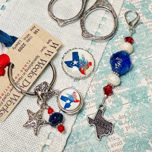 May include: A red, white, and blue themed charm with a silver key ring, a star charm, and a Texas-shaped charm. The charm also includes a blue and white bead, a red and white bead, and a blue bead. The charm is hanging from a tag that says "WW Weeks Dy Works" and "Specializing in Hand Dyed Fibers".