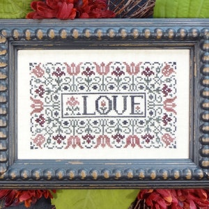 Puede incluir: Un bordado de punto de cruz enmarcado con un diseño floral y la palabra "LOVE" en el centro. El marco es negro con un borde de cuentas.
