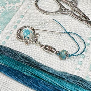 May include: A silver needle threader with a blue and white turtle charm, a blue and green glass bead, and a length of blue embroidery floss.