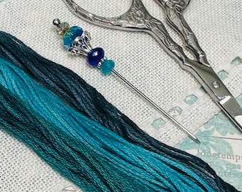 May include: A close-up of embroidery supplies, including a silver needle with blue and teal beads, silver scissors, and skeins of teal and navy blue embroidery floss. The needle is placed next to the scissors and floss on a white fabric.