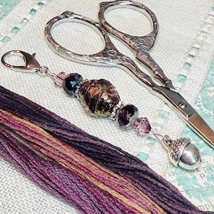 May include: An image of embroidery tools: silver scissors with decorative handles, a beaded charm with an acorn pendant, and skeins of purple, pink, and gold embroidery thread. The items are on a textured surface.