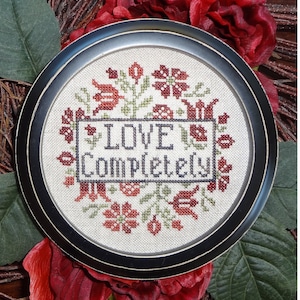 Puede incluir: Un bordado a punto de cruz enmarcado con fondo blanco y bordado floral en rojo y verde. El bordado dice "LOVE COMPLETELY".