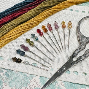 May include: A set of ten colourful glass-headed pins with silver metal shafts, a pair of silver embroidery scissors, and several spools of embroidery floss in shades of green, brown, and yellow.