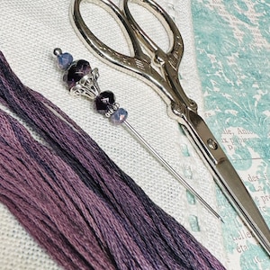 May include: A silver needle threader with a purple and clear glass bead embellishment. The needle threader is lying on a white fabric surface with a pair of silver scissors and purple embroidery floss.