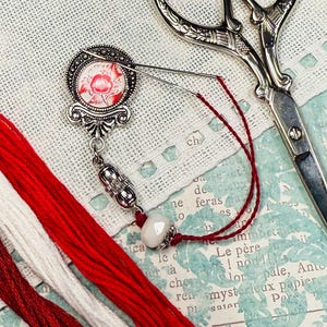 May include: A silver needle minder with a floral design, red embroidery floss, and vintage scissors. The needle minder features a decorative pendant with a red flower image. The scissors are silver.