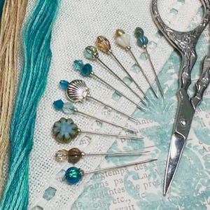 May include: A selection of decorative sewing pins with bead embellishments, including a seashell, alongside teal and beige embroidery threads. A pair of decorative silver scissors is also visible, indicating a crafting project.