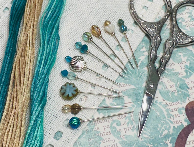 May include: A selection of decorative sewing pins with bead embellishments, including a seashell, alongside teal and beige embroidery threads. A pair of decorative silver scissors is also visible, indicating a crafting project.