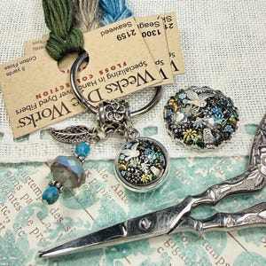 May include: A collection of craft supplies including embroidery floss, a charm with a bird design, a decorative button, and ornate scissors. The floss is labeled "Weeks Dye Works". The charm and button feature a floral and bird motif.
