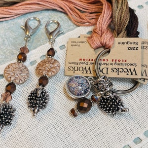 May include: A silver key chain with a variety of charms, including a floral design, a paisley pattern, and a spiky ball. The key chain is attached to a skein of embroidery floss in shades of brown, pink, and beige. The text on the card reads "Weeks Dy Works", "Specializing in Hand Dyed Fibers", "FLOSS COLLECTION", "123", "2253", "Flatf Sanguine", and "5 yards Cotton Floss".