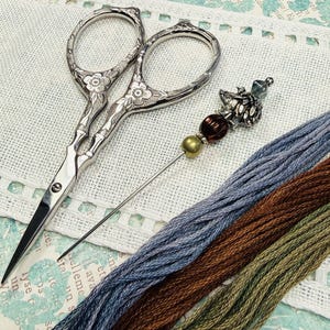 May include: Silver embroidery scissors with floral detailing, a decorative needle, and three skeins of thread in blue, brown, and green. The scissors and needle rest on a white fabric with a decorative edge.