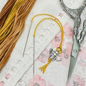 May include: A close-up of a silver bee charm with a yellow bead, attached to a length of yellow embroidery floss. Two embroidery needles are also visible, along with a pair of silver scissors and a piece of white lace fabric.