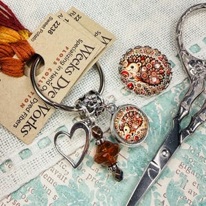 May include: A selection of craft items featuring Weeks Dye Works floss, a heart charm, a hedgehog-themed button, a hedgehog pendant, and decorative scissors. The floss is labelled "Sweet Potato".
