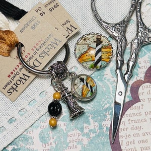 May include: A silver key ring with a lighthouse charm, a map charm, and a set of beads. The beads are black, gold, and white. The key ring is on a white fabric background.