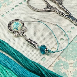 May include: A silver needle minder featuring a circular cabochon with a teal turtle graphic, connected to a cylindrical silver magnet and a single faceted teal bead, with a sewing needle attached and teal embroidery floss strands nearby.