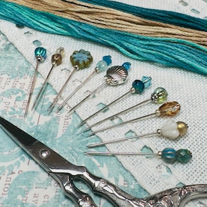 May include: A selection of decorative pins with diverse bead and charm designs, including a seashell, and colourful beads. The pins are arranged with embroidery floss in shades of teal and tan, and a pair of silver scissors.