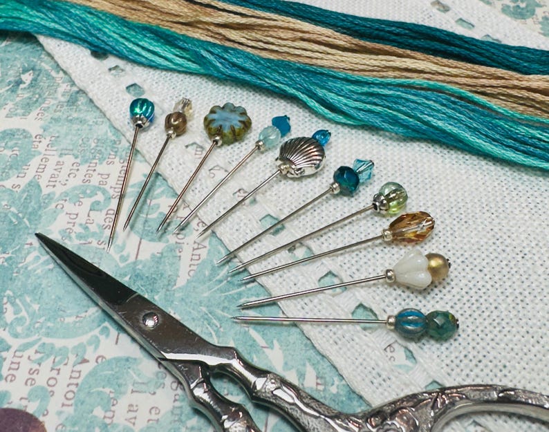 May include: A selection of decorative pins with diverse bead and charm designs, including a seashell, and colourful beads. The pins are arranged with embroidery floss in shades of teal and tan, and a pair of silver scissors.